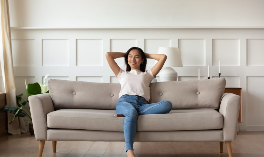 Asian woman looks at distance resting leaned on couch enjoy fresh air in summer day in modern fashionable living room interior, full length image. Contemporary apartments owner or carefree day concept