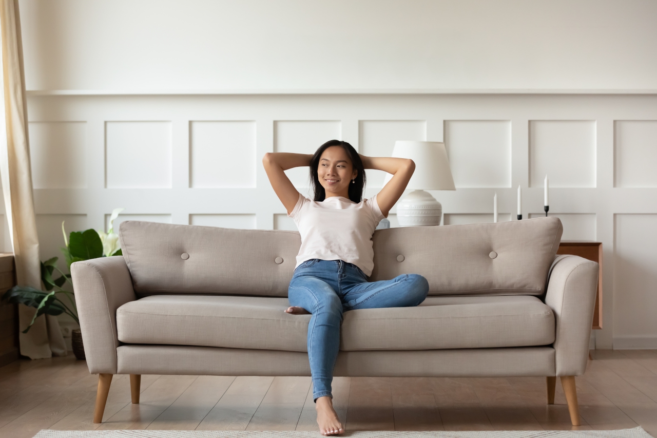 Asian woman looks at distance resting leaned on couch enjoy fresh air in summer day in modern fashionable living room interior, full length image. Contemporary apartments owner or carefree day concept