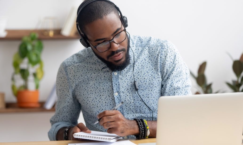 Focused african business man in headphones writing notes in notebook watching webinar video course, serious black male student looking at laptop listening lecture study online on computer e learning