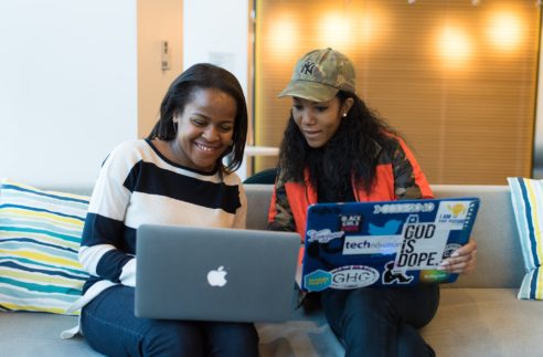 two black women chatting on a couch with their laptops
