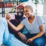 Cheerful dark skinned couple in love resting at home interior posing for selfie on smartphone camera,happy young hipsters making image on modern mobile phone laughing at living room together