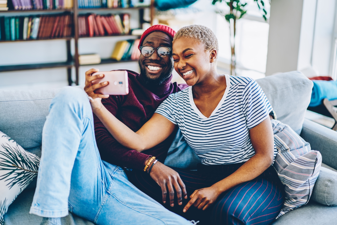 Cheerful dark skinned couple in love resting at home interior posing for selfie on smartphone camera,happy young hipsters making image on modern mobile phone laughing at living room together
