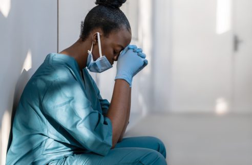 Tired depressed female african scrub nurse wears face mask blue uniform gloves sits on hospital floor. Exhausted sad black doctor feels burnout stress of corona virus frontline protection pray at work