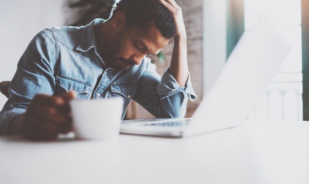 Tired young African man using laptop while sitting at the table on a sunny morning.Concept of people working hard home.Blurred background,flare effect