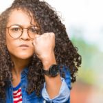 Young beautiful woman with curly hair wearing glasses angry and mad raising fist frustrated and furious while shouting with anger. Rage and aggressive concept.
