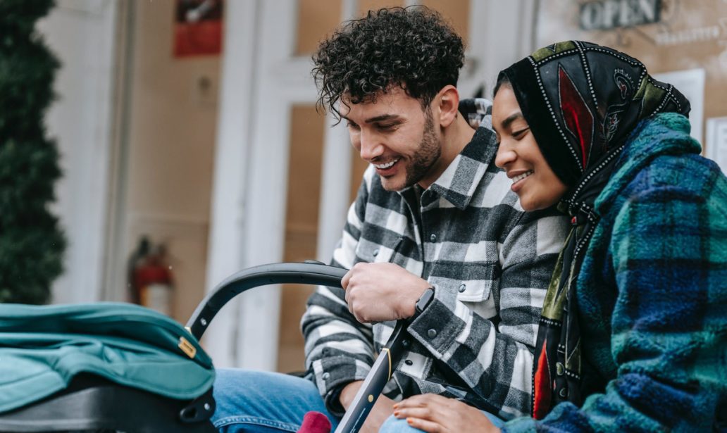 cheerful young multiethnic spouses smiling and looking at baby in stroller