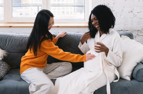 happy asian woman touching belly of pregnant black partner