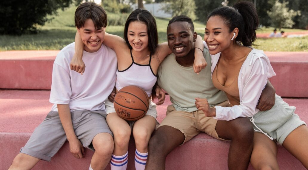 happy multiracial friends embracing on bench after playing basketball