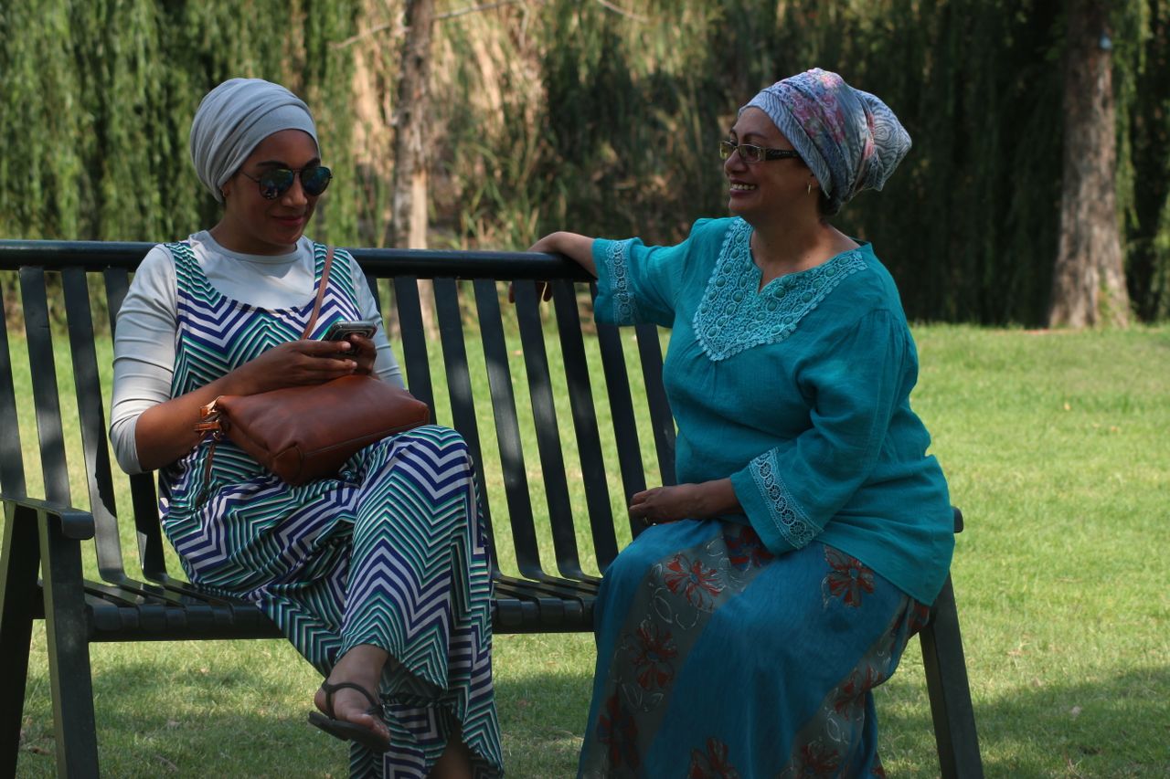Two women in colorful attire enjoy leisure time on a bench in a sunny park.
